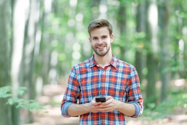 photo of positive man messaging on phone in the forest. man messaging on phone outdoor. man messaging on phone outside. man messaging on phone wearing checkered shirt.