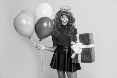 Sarı arka planda hediye kutusu ve parti balonuyla taç giymiş mutlu sürpriz çocuk..