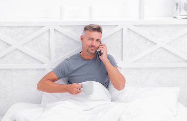 Sabah çayı ya da yatakta kahve içerken cep telefonuyla konuşan mutlu adam, ara..