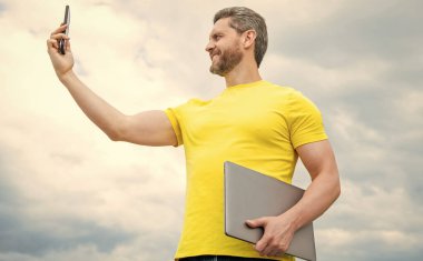 man with laptop making selfie on smartphone on sky background.