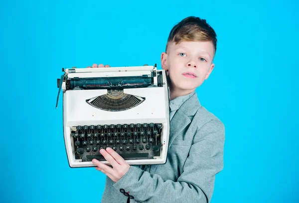 Using retro technology. Little boy holding retro typewriter on blue background. Small boy having old typewriter. Smart boy with vintage typewriter. Cute boy with mechanical desktop typewriter.