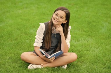 Gözlüklü çocuk yeşil çimlerin üzerinde oturmuş kitap okuyor..
