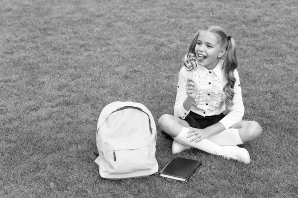 スクールバッグと制服を着た幸せな女の子の子供と本は緑の芝生の上に座っておいしいロリポップを食べることを楽しむ — ストック写真