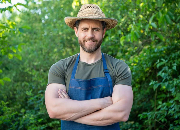 Happy gardener man smiling in gardening apron and farmers hat keeping arms crossed in garden outdoors.