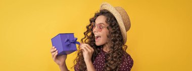 Kıvırcık saçlı hayret bir çocuk sarı arka planda hediye kutusunu tutuyor..