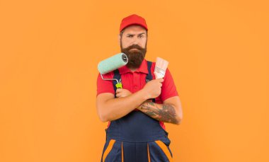 serious bearded man decorator in work clothes hold paint roller and brush on yellow background.