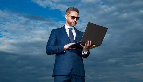 空を背景にノートパソコンを入力するビジネスマン ノートパソコンの屋外で ノートパソコンのスーツを着たビジネスマン ノートパソコンを持ったビジネスマンの写真 — ストック写真