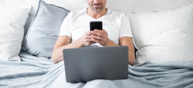 Neşeli, olgun bir adam telefonda konuşuyor ve laptopla yatakta çalışıyor..