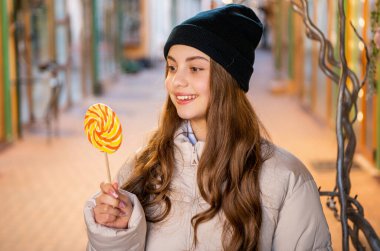 Lolipop şekerli, şapkalı gülümseyen kız. Dışarıda lolipop şekerli bir kızın fotoğrafı var. Sokakta elinde lolipop şekeri olan kız. Açık havada lolipop şekerli kız.