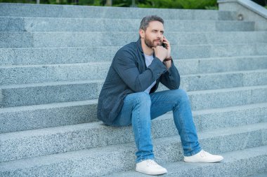 Telefondaki adam dışarıda oturuyor. Adamın telefonla arayıp konuştuğu bir fotoğraf. Adamın dışarıda telefonu var. Sokakta telefonla konuşan bir adam var..