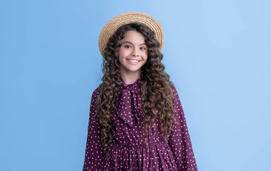 child smile in straw hat with long brunette curly hair on blue background.