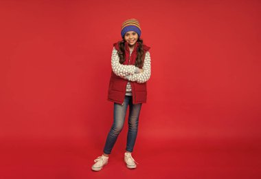 Kış şapkası takmış, kırmızı arka planda yelek giyen, kışın giyinen bir çocuk..