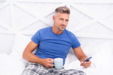 Yatakta kahve içerken cep telefonuyla sosyal medyada sörf yapan boz saçlı adam. Mobil cihazda mobil internet kullanılıyor. Mobil iletişim.