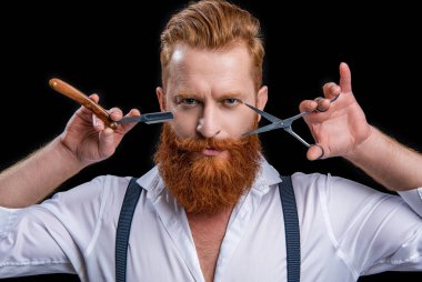 Makas ve jiletle traşsız berber. Arka planda berberin fotoğrafı var. Berber adam siyahlar içinde izole edilmiş. Stüdyoda berber..