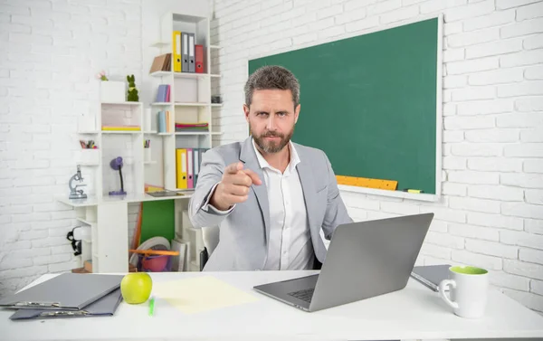 serious school teacher in classroom with computer at blackboard. point finger.