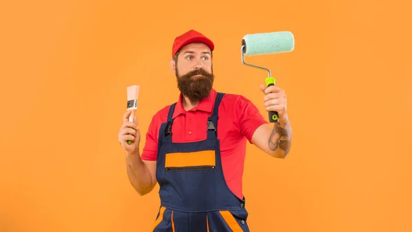 puzzled bearded man in work clothes hold paint roller and brush on yellow background.
