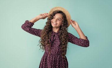 cheerful kid in straw hat with long brunette curly hair on blue background.