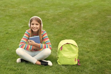 Happy teenage girl listening to audio training course in headphones sitting on grass, school.