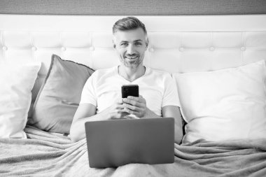 positive mature man chatting on phone and working on laptop in bed.