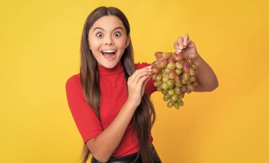 Afallamış genç kız gülümsüyor sarı arka planda bir demet üzüm tutuyor.
