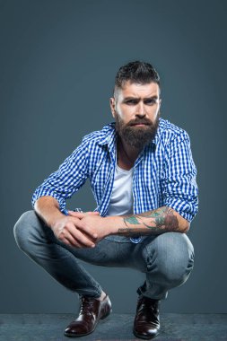 Sakallı adam gri arka planda oturan kareli gömlek giyiyor..