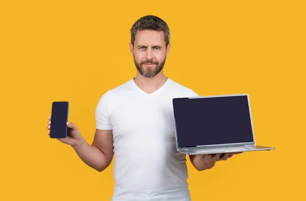 guy showing smartphone and laptop screen in studio. guy showing screen of laptop and smartphone. guy showing screen with copy space. guy showing smartphone screen isolated on yellow background.