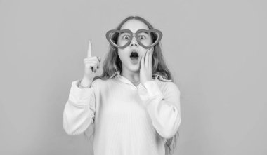 Surprised kid in funny heart-shaped glasses keeping finger raised blue background, eureka.