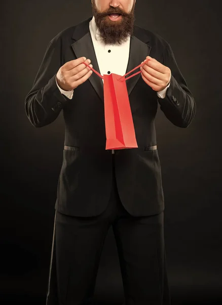 surprised curious man in tuxedo bow tie formalwear on black background with business reward in shopping bag. occasion greeting.