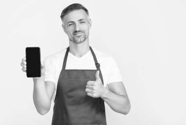 cheerful mature man in apron presenting smartphone with copy space. thumb up.