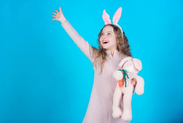 Child smiling play bunny toy. Happy childhood. Get in easter spirit. Bunny ears accessory. Lovely playful bunny child with long hair. Cute and adorable. Bunny girl with cute toy on blue background.
