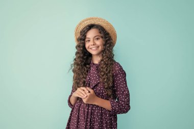 smiling child in straw hat with long brunette curly hair on blue background.