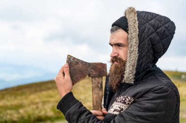 Keskin baltası olan sakallı bir oduncu. Dağda balta tutan sakallı bir oduncunun fotoğrafı. Sakallı adam dışarıda baltayı tutuyor. Sakallı adamın baltası var..