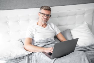 mature man virtual assistant in glasses working on laptop in bed.
