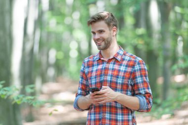 Adam dışarıda telefonda gülümsüyor ve mesajlaşıyor. Adam dışarıda telefonla mesajlaşıyor. Telefondaki mesajlaşan adam kareli gömlek giyiyor. Ormanda telefonla mesajlaşan bir adamın fotoğrafı..