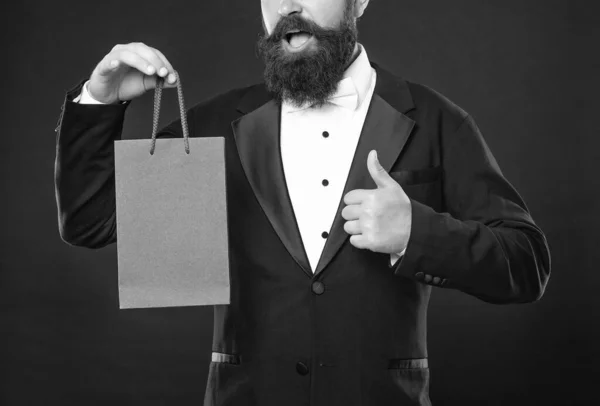 amazed man in tuxedo bow tie formalwear on black background with purchase shopping bag. copy space. thumb up.