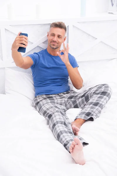 cheerful blogging man with phone in bed. photo of blogging man with phone at home. blogging man with phone in bedroom. blogging man with phone wear pajama.