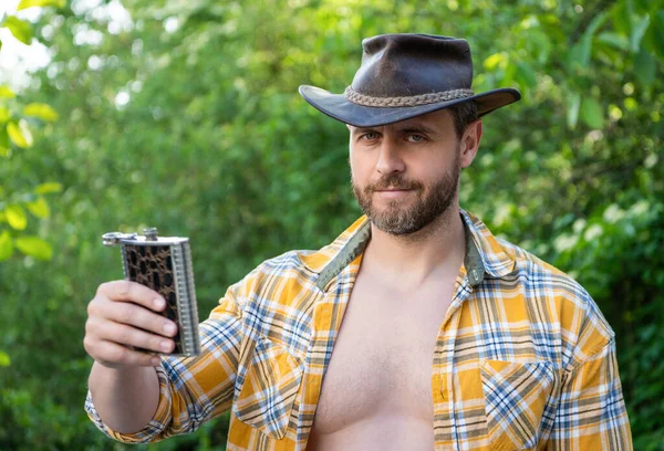 photo of western man offering whiskey flask. western man with whiskey flask. western man with whiskey flask outdoor. western man with whiskey flask wear checkered shirt.