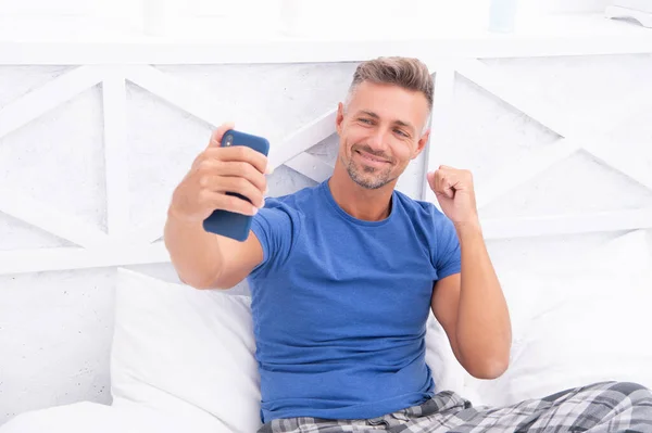 photo of successful blogging man with phone at home. blogging man with phone in bedroom. blogging man with phone wear shirt. blogging man with phone in bed.