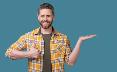 Olgun bir adam damalı gömlek giyer, ürünleri sunar ve başparmağını kaldırır. Sakallı, olgun bir adamın fotoğrafı. Olgun bir adam mavi arka planda izole edilmiş. Stüdyoda olgun bir adam.