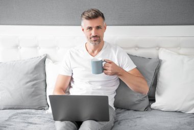 mature man smile using computer in bed with coffee.