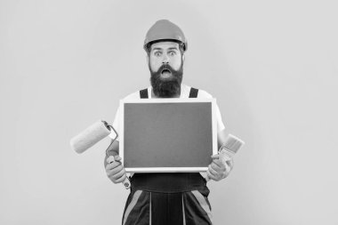 shocked man renovator in helmet and work clothes hold paint roller brush and blackboard with copy space on yellow background.