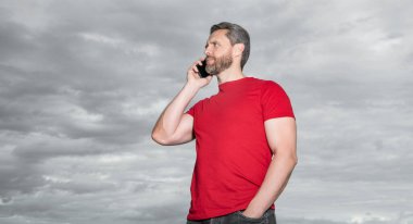 caucasian man has phone talk outdoor. man has phone talk wear red tshirt. photo of man has phone talk. man has phone talk on sky background.