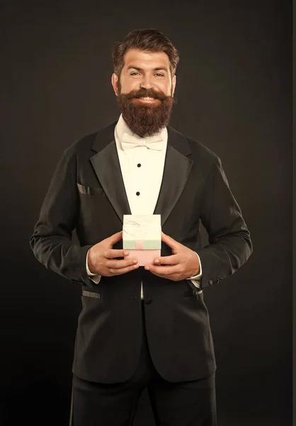 cheerful man in tuxedo bow tie formalwear on black background with business reward box. occasion greeting.