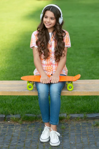 スケートボードの屋外で幸せな10代の女の子 ヘッドフォンでスケートボードを持つ10代の女の子 スケートボードを持ってる10代の女の子 スケートボードで10代の女の子の写真は音楽を聞く — ストック写真