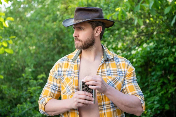 western man with whiskey flask outdoor. western man with whiskey flask wear checkered shirt. photo of western man with whiskey flask. western man with whiskey flask.