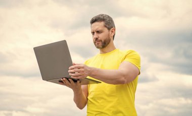 Gökyüzü arka planında dizüstü bilgisayarda çalışan konsantre bir adam.