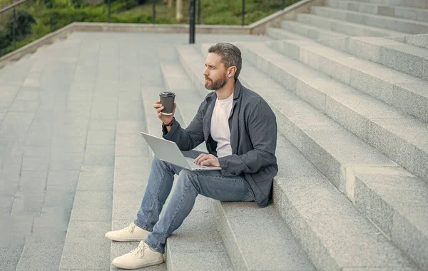 stock image photo of man blogging online with computer having coffee. man blogging online with laptop outdoor. man blogging online in the street. man blogging online with pc outside.