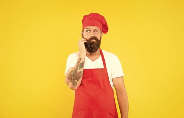 Hesitant man in red toque and apron stroking chin yellow background, chef.