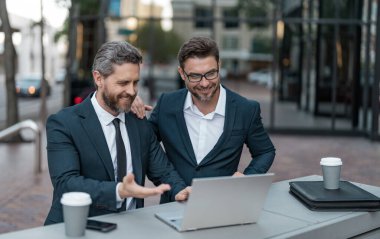 Neşeli iş adamları bilgisayarla çevrimiçi ticaret yapıyor. İnternet üzerinden ticaret yapan iş adamlarının fotoğrafı. İki işadamı çevrimiçi ticaret yapıyor. İş adamları açık havada ticaret yapıyor.