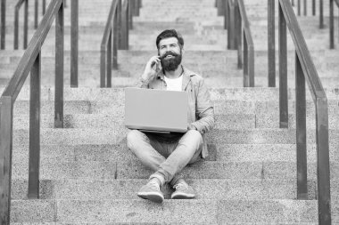 glad guy has online video communication sitting on stairs. guy has online video communication on laptop. guy has online video communication outdoor. guy has online video communication outside.